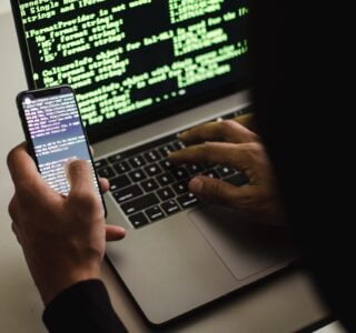 Free Unrecognizable hacker with smartphone typing on laptop at desk Stock Photo