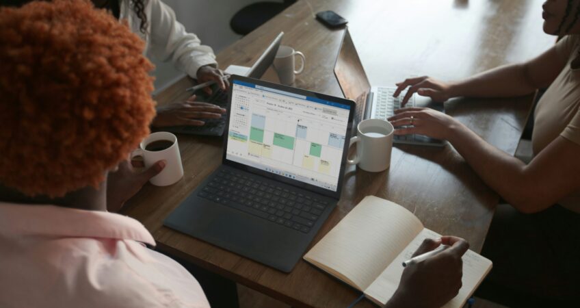 Three people in a meeting at a table discussing schedule on their Microsoft laptop